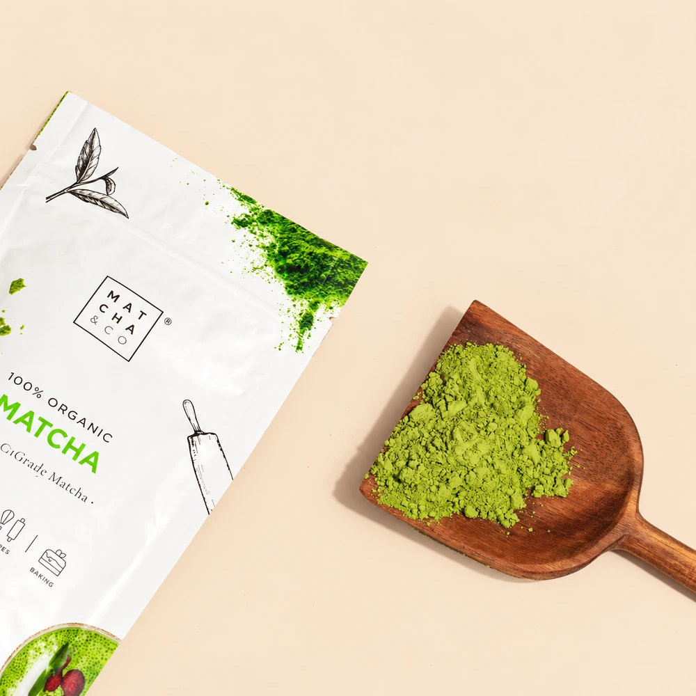 Πράσινη σκόνη matcha σε ξύλινη κουτάλα δίπλα σε συσκευασία Matcha & Co.