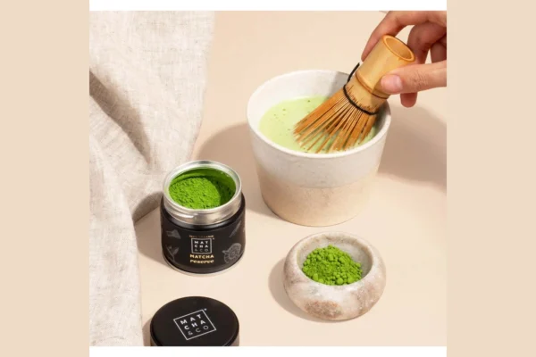 Παρασκευή matcha με χτυπητήρι μπαμπού σε κεραμικό μπολ, δίπλα σε συσκευασία Matcha & Co Reserve.